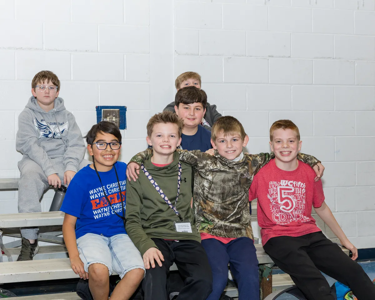 Seven boys sit together on gym bleachers posing for a photo with a white brick wall behind them.